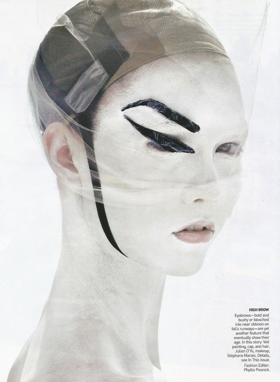 A woman with white face paint, dramatic black eye makeup, and a sheer head covering poses in a studio, creating an avant-garde fashion look.