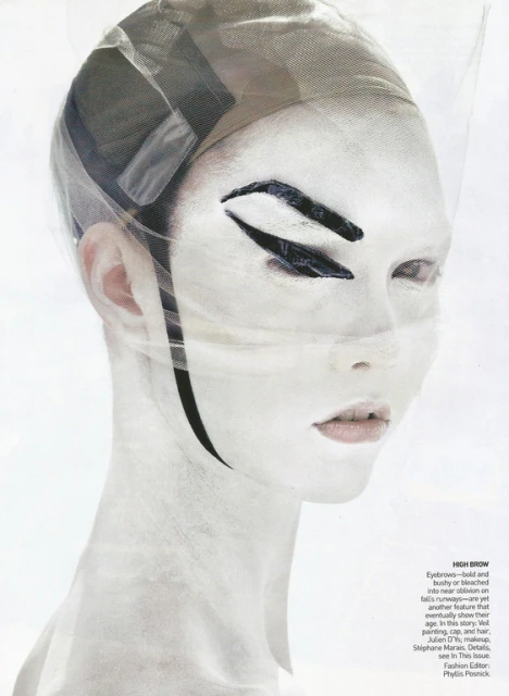 A woman with white face paint, dramatic black eye makeup, and a sheer head covering poses in a studio, creating an avant-garde fashion look.