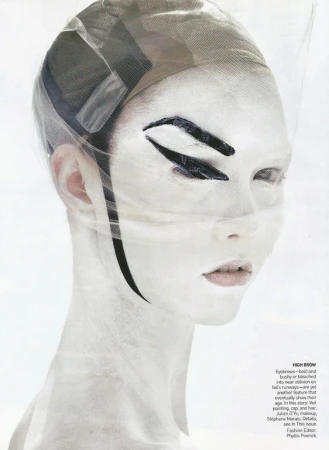 A woman with white face paint, dramatic black eye makeup, and a sheer head covering poses in a studio, creating an avant-garde fashion look.