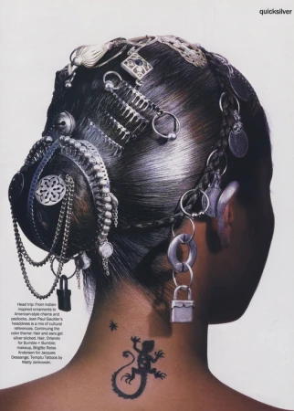 Woman's head and shoulders shot from behind, intricate hairstyle with ornamental hair accessories and neck tattoo detail against a white backdrop.