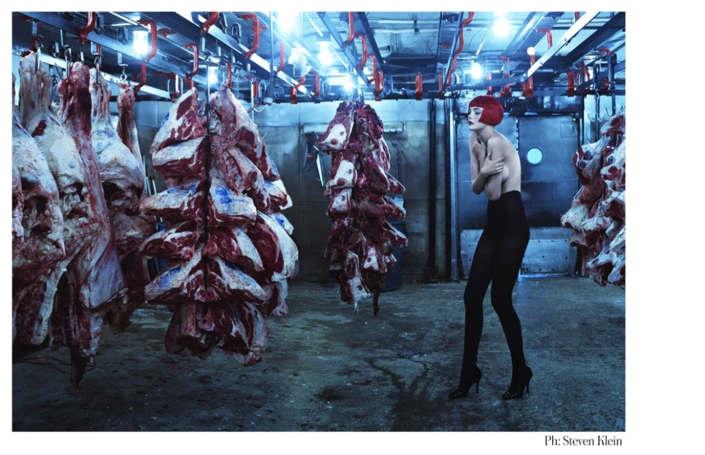 A bare-shouldered adult female with red hair poses among sides of beef hanging in a cold storage warehouse or meat-packing facility.