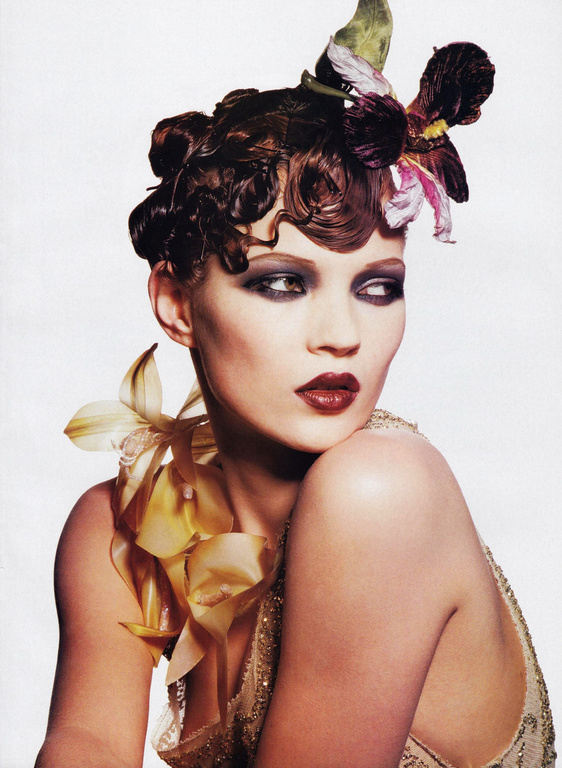 Close-up of an adult woman wearing dark eye makeup and lipstick, adorned with a floral headpiece and flower around the neck.