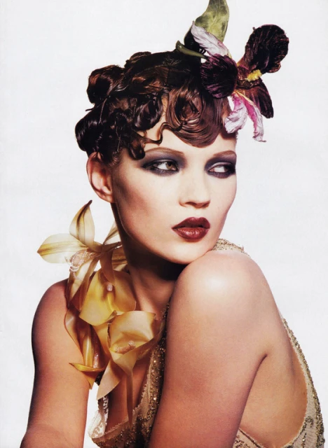 Close-up of an adult woman wearing dark eye makeup and lipstick, adorned with a floral headpiece and flower around the neck.
