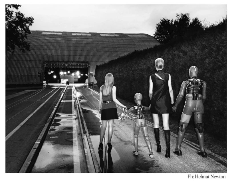 Black and white image of two women and two mannequins walking on a wet road, with a building with arched entrance in the background.
