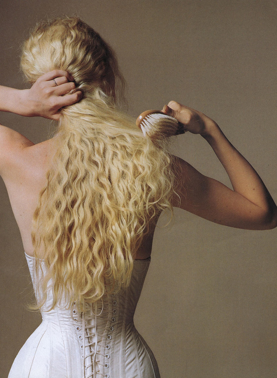 Woman brushing long blonde wavy hair while wearing a white corset top, shown from the back against a neutral background.