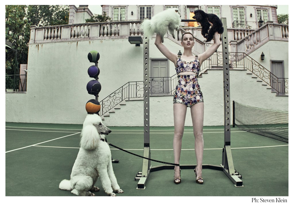 Adult female in a floral outfit holds a white dog and a black dog above her head next to a weight rack on a tennis court.