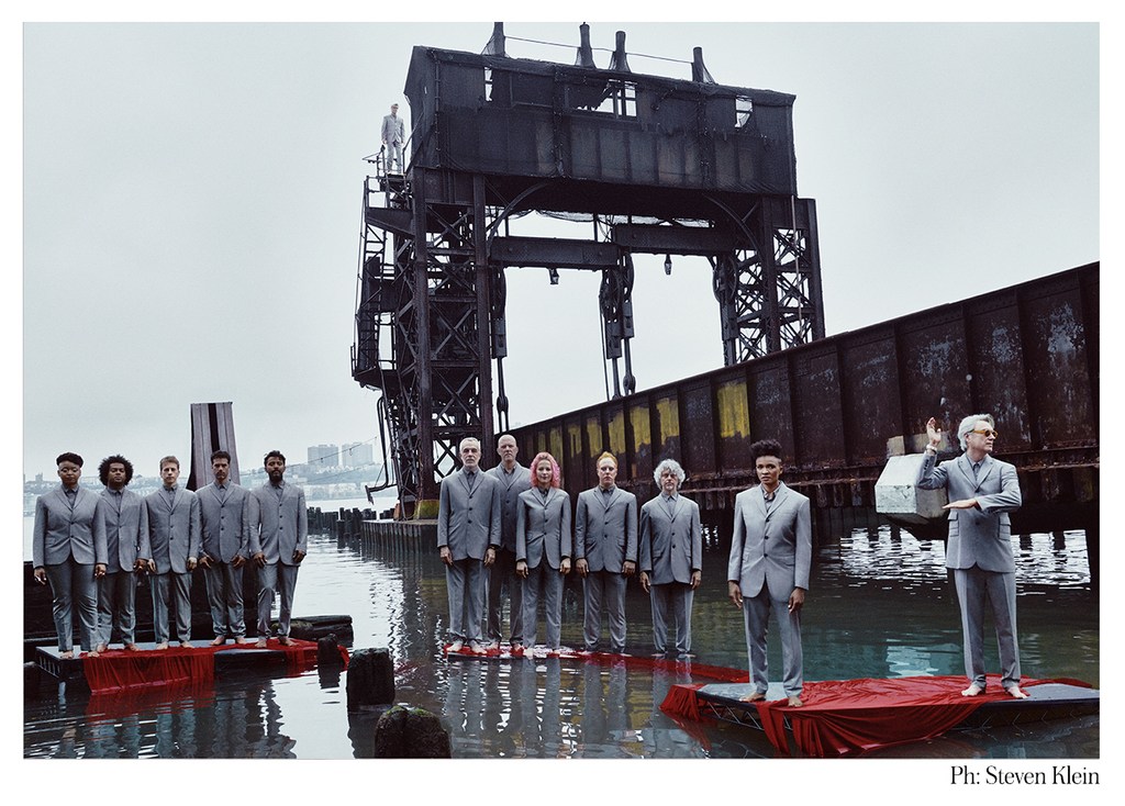 A group of people dressed in suits standing on platforms in water with an industrial structure and overcast sky in a stylized editorial.