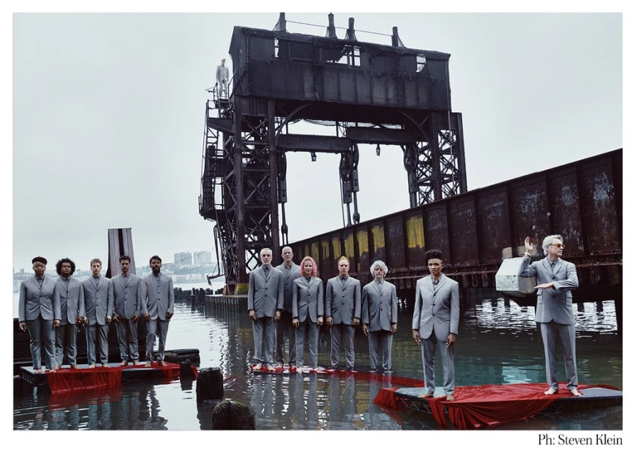 A group of people dressed in suits standing on platforms in water with an industrial structure and overcast sky in a stylized editorial.