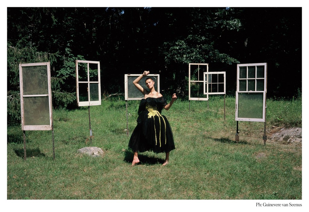 Woman in a black dress poses barefoot in a grassy field surrounded by antique window frames on poles in an outdoor setting.