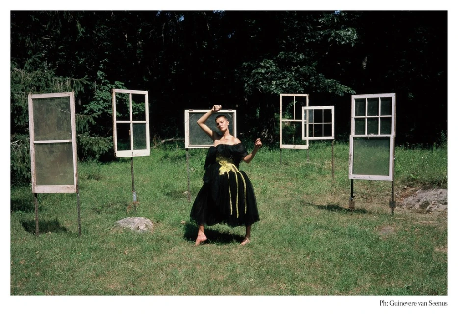 Woman in a black dress poses barefoot in a grassy field surrounded by antique window frames on poles in an outdoor setting.