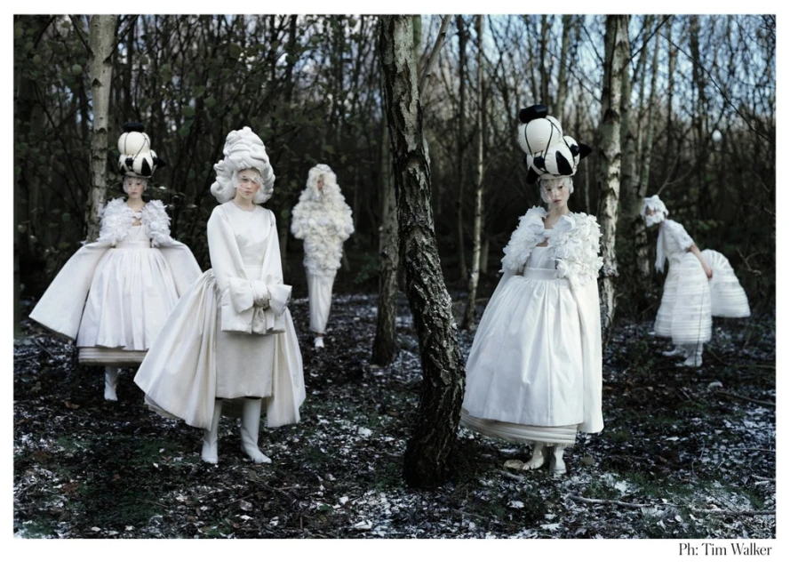 A group of five women in elaborate white dresses and headwear stand in a dark forest, posing for a high-fashion editorial photoshoot.