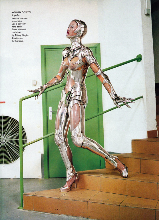 Fashion editorial shot of a slender adult female in a silver metallic robot suit posing on stairs with a green door and AC unit in the background.