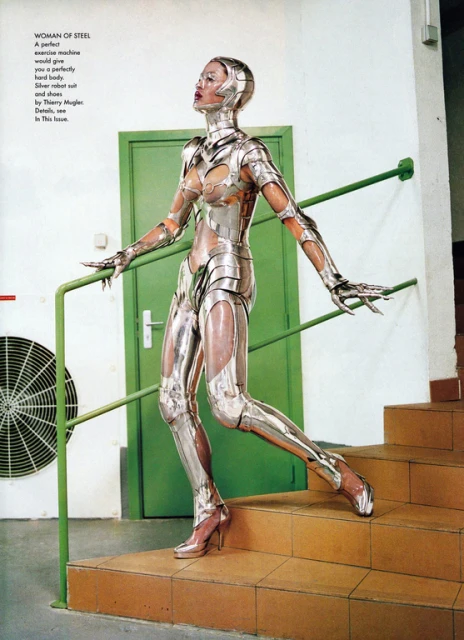 Fashion editorial shot of a slender adult female in a silver metallic robot suit posing on stairs with a green door and AC unit in the background.