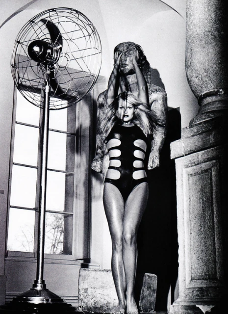 Black and white studio shot of a woman in a black bodysuit standing in front of a statue and a metal fan, creating a stark, staged scene.