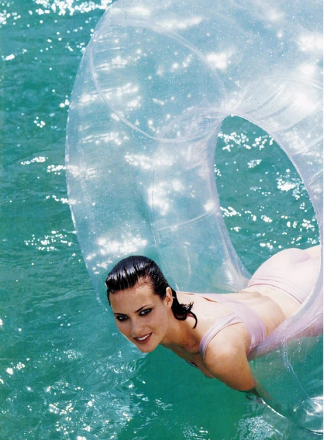 Woman with wet hair is lying in a clear inflatable pool toy on shimmering water in an eye-level shot with medium framing and pale colors.