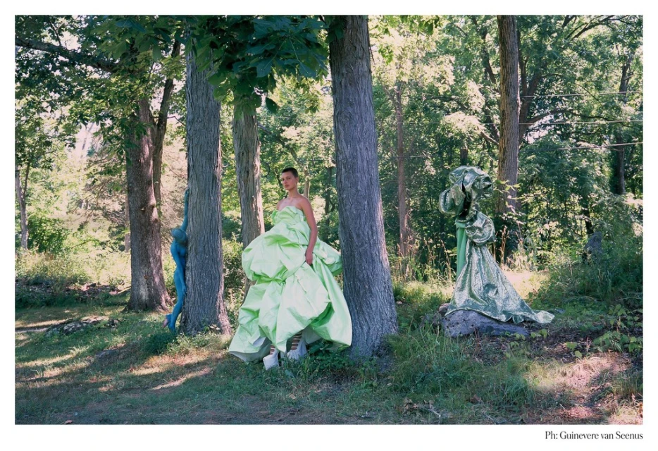 Woman in an apple-green ball gown stands in the woods. Sculptures sit on the ground. Trees provide a backdrop for the outdoor setting.
