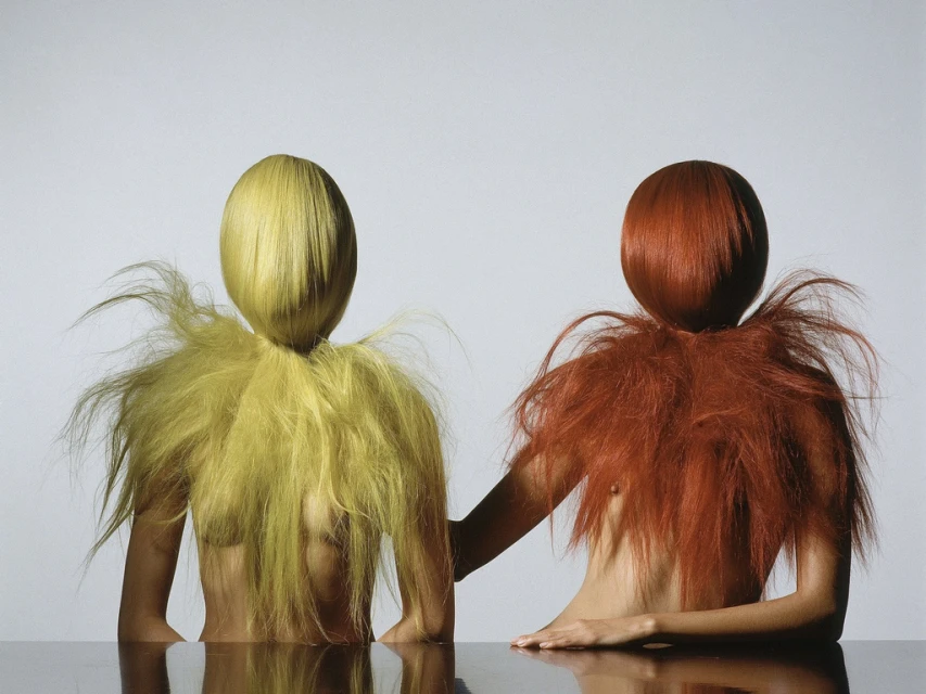 Two models with yellow and red hair and fur-like collars pose at a shiny table in a studio, creating a surreal and artistic portrait.