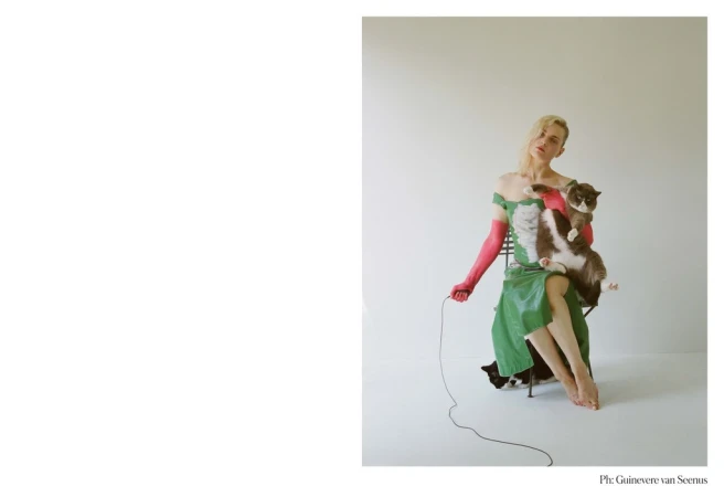 Woman in a green dress sits on a chair holding a fluffy grey and white cat, posing in front of a neutral backdrop with simple lighting.