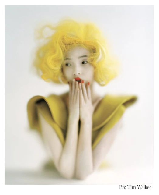 Portrait of an Asian woman with yellow, voluminous, curly hair. Her hands are up to her face, and she is wearing red lipstick.