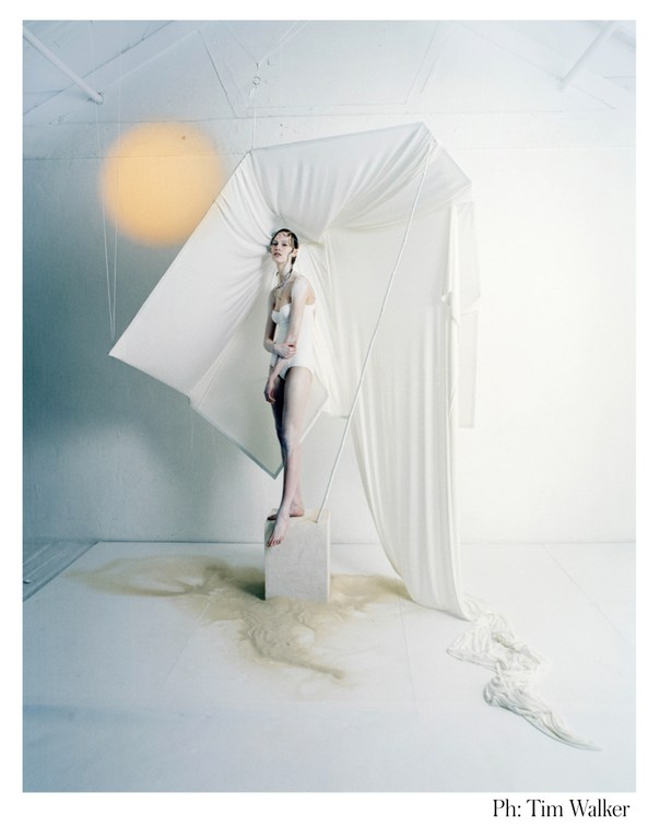 Artistic fashion image in a studio featuring a woman in white lingerie standing on a block under a draped cloth structure.