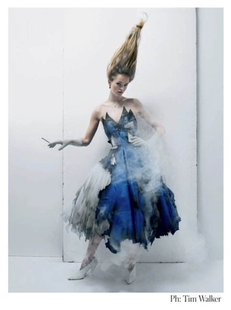 A woman with blonde hair standing in a studio setting. She is wearing a blue and white dress with a cloud-like design and white heeled shoes.