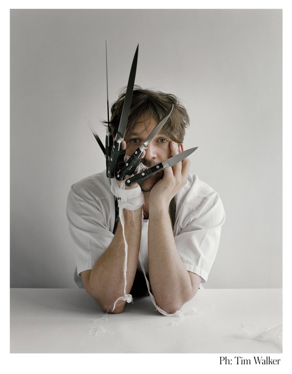 Portrait of a man with a knife-blade glove on one hand, resting his face in his hands, looking directly at the camera against a grey backdrop.