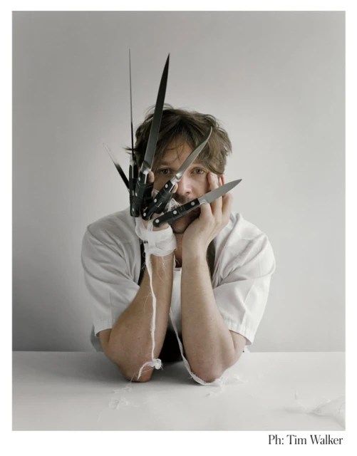 Portrait of a man with a knife-blade glove on one hand, resting his face in his hands, looking directly at the camera against a grey backdrop.