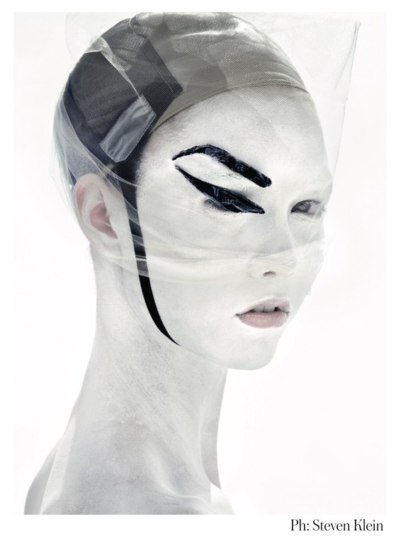 Artistic fashion portrait of a woman with white face paint and bold eye makeup in a bright studio environment.
