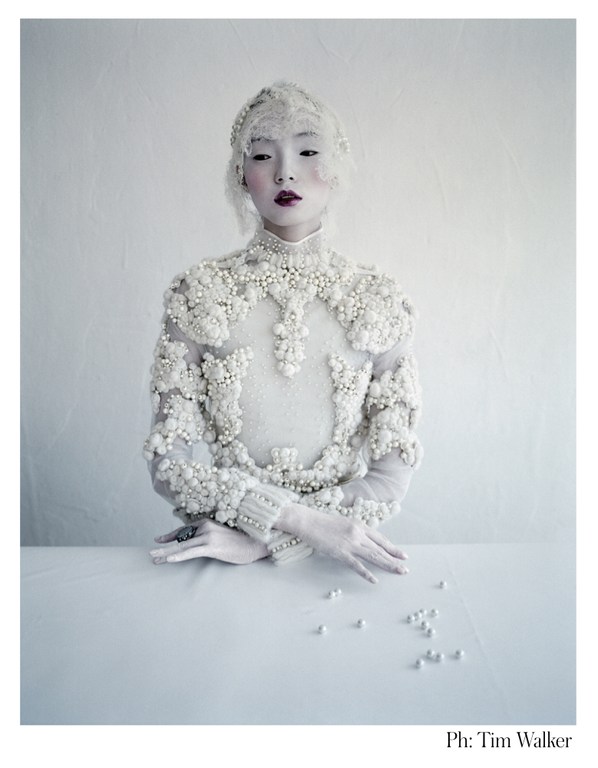 Elegant young Asian woman in a white gown and headpiece at a white table, adorned with pearls, captured in a studio shot.