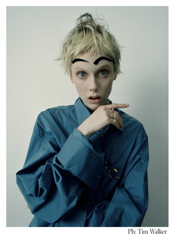 Blonde woman poses with exaggerated eyebrows in a fashion shot, wearing a blue shirt and gold rings against a white backdrop.
