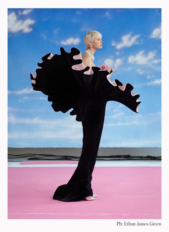 Fashion model in black ruffled gown strikes pose against vibrant blue sky with clouds and pink floor for editorial fashion portrait.