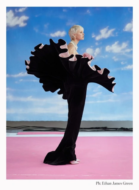 Fashion model in black ruffled gown strikes pose against vibrant blue sky with clouds and pink floor for editorial fashion portrait.
