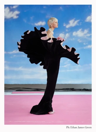 Fashion model in black ruffled gown strikes pose against vibrant blue sky with clouds and pink floor for editorial fashion portrait.