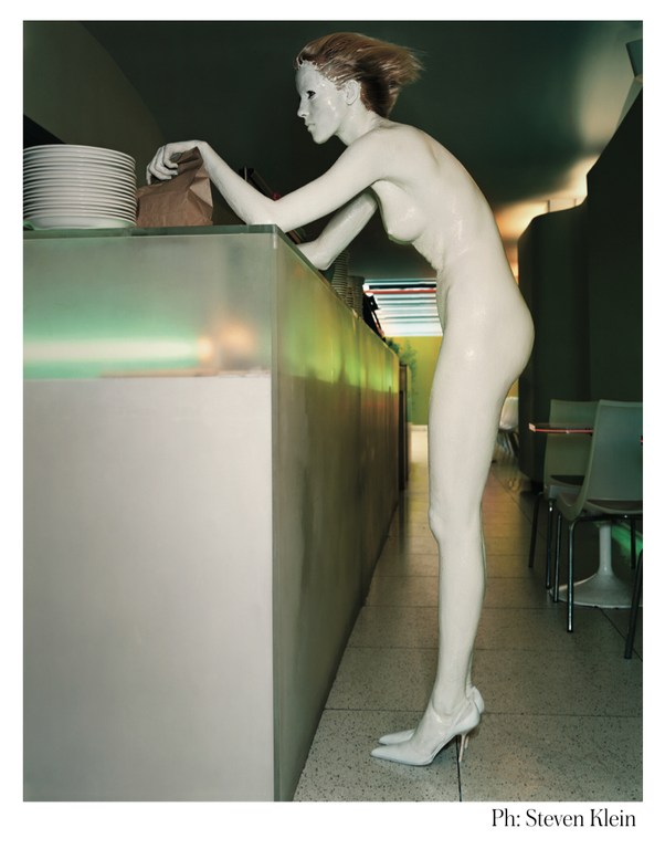 Full-body studio shot of a light-skinned mannequin in high heels, leaning on a counter with plates, in a dimly lit interior space.