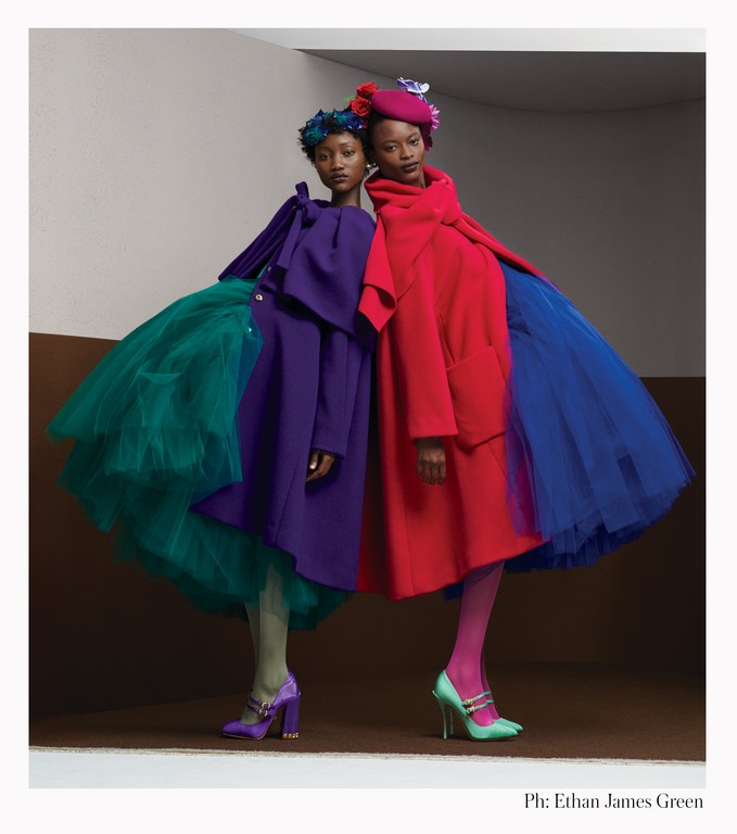 Two fashion models pose in vibrant, oversized coats with tulle skirts, floral headpieces, and high heels in a studio setting.