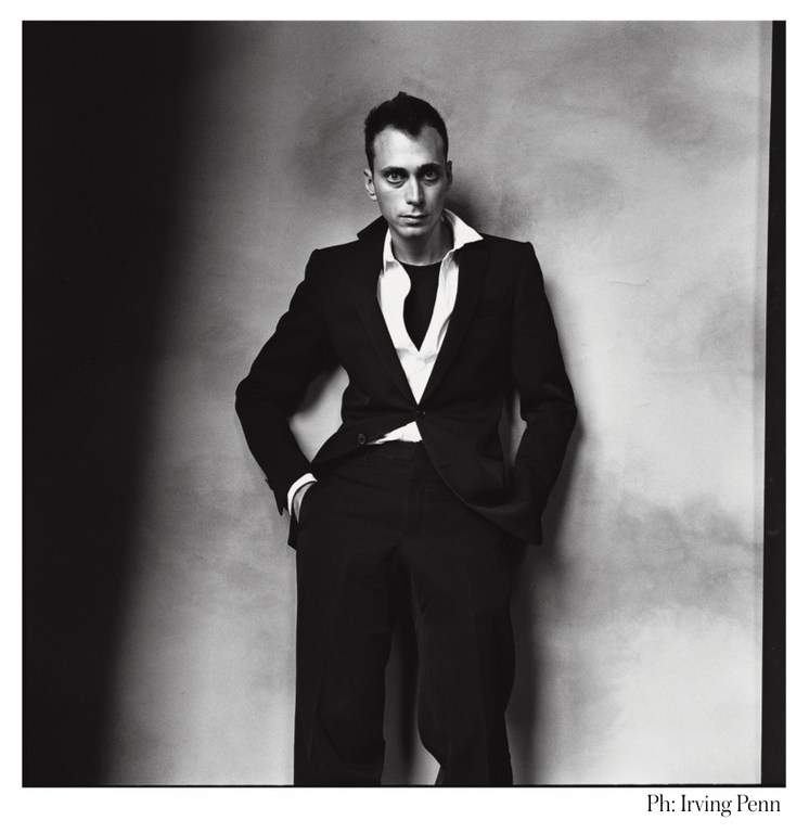 Black and white portrait of a man in a dark suit and white shirt, standing against a mottled gray wall with dark shadows.