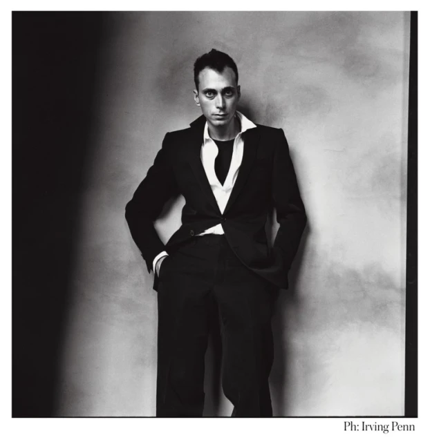 Black and white portrait of a man in a dark suit and white shirt, standing against a mottled gray wall with dark shadows.