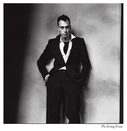 Black and white portrait of a man in a dark suit and white shirt, standing against a mottled gray wall with dark shadows.
