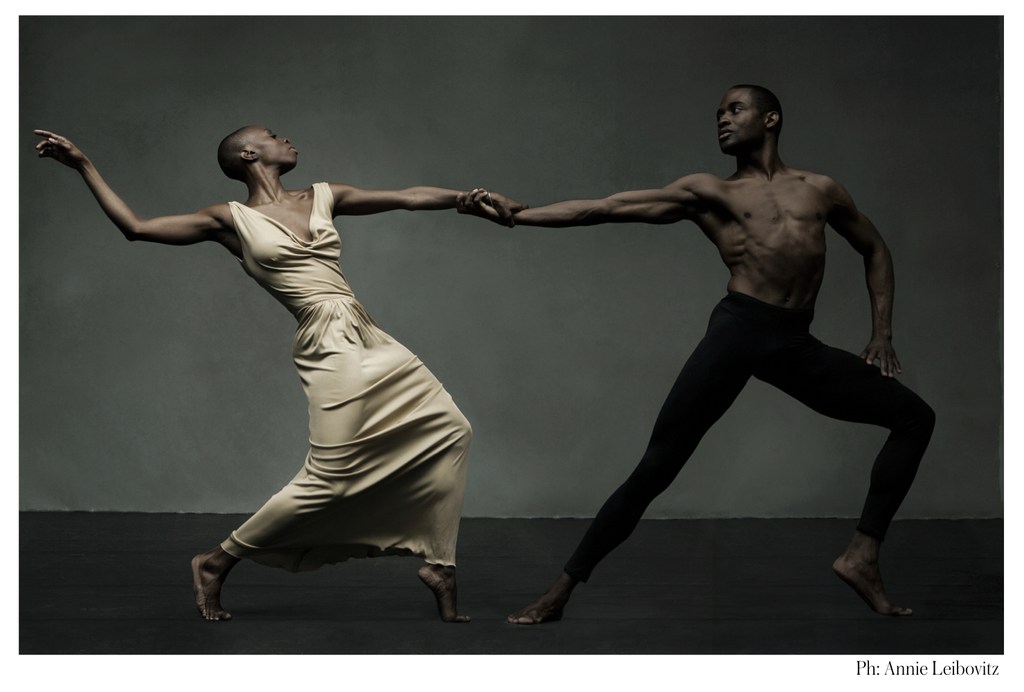 Two dancers, an adult male and adult female, hold hands while posing in a dance move in front of a neutral gray background.