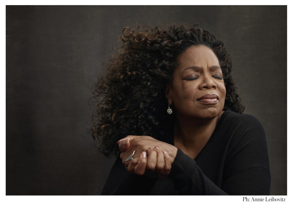 Portrait of Oprah Winfrey with closed eyes, wearing a black top and jewelry, against a textured dark grey background in studio lighting.