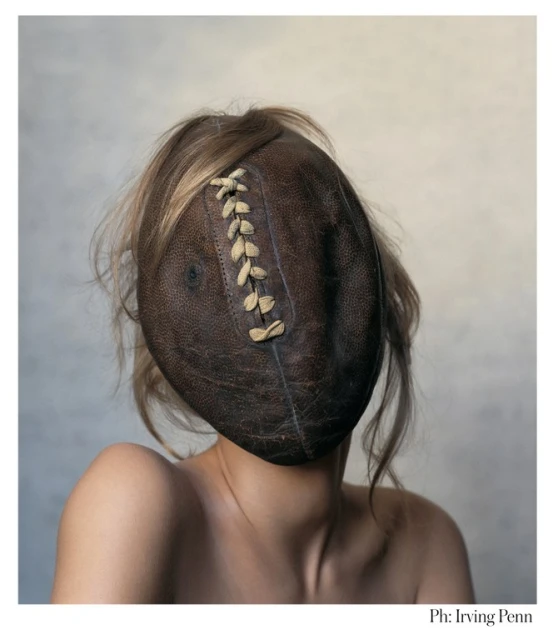 Conceptual portrait of a topless adult female with blond hair, her face obscured by a brown leather American football.