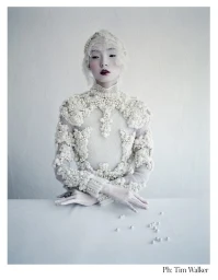 Model with white face paint wearing an elaborate beaded white top and lace headpiece, seated behind loose pearls.