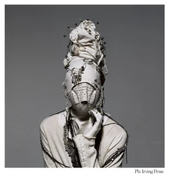 Avant-garde portrait of a model wearing a white fabric headpiece and face covering secured with numerous safety pins, styled by Phyllis Posnick.