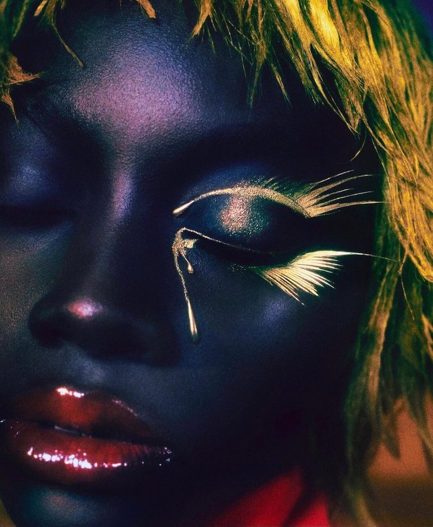 Close-up of a woman's face featuring gold artistic makeup, closed eyes, and glossy red lips against a dark backdrop in a beauty shot.