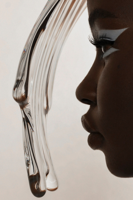 Close-up studio shot of a dark-skinned woman in profile with white makeup, posing near a textured glass prop against a neutral background.