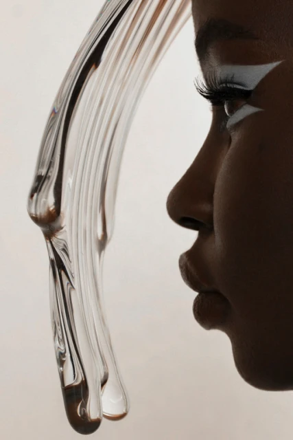 Close-up studio shot of a dark-skinned woman in profile with white makeup, posing near a textured glass prop against a neutral background.