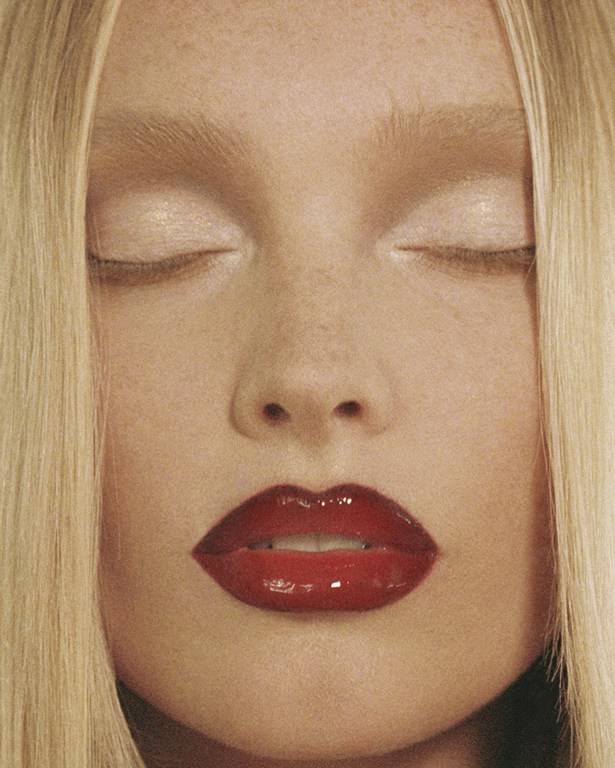 Close-up portrait of a fair-skinned woman with blonde hair and red lips, eyes closed, showcasing beauty and makeup in a studio setting.