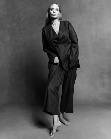 Monochrome portrait of Angelina Jolie wearing a dark oversized double-breasted suit and trousers, standing barefoot on a studio backdrop.