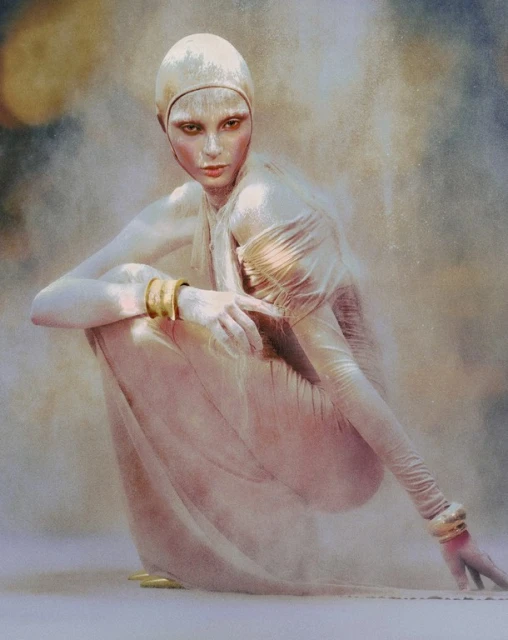 Crouching woman in pale gown and swim cap poses artistically against a neutral background, adorned with gold bracelets and ankle cuffs.