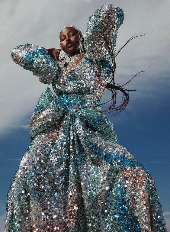 Woman in a shimmering turquoise and silver gown poses against a blue sky, looking up and away in a fashion editorial photograph.